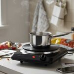 OVENTE 1000W single burner hot plate with cast iron surface heating a small pot on a cluttered apartment counter