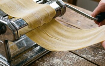 Hands adjusting the dial on a stainless steel pasta maker on a flour-dusted counter.