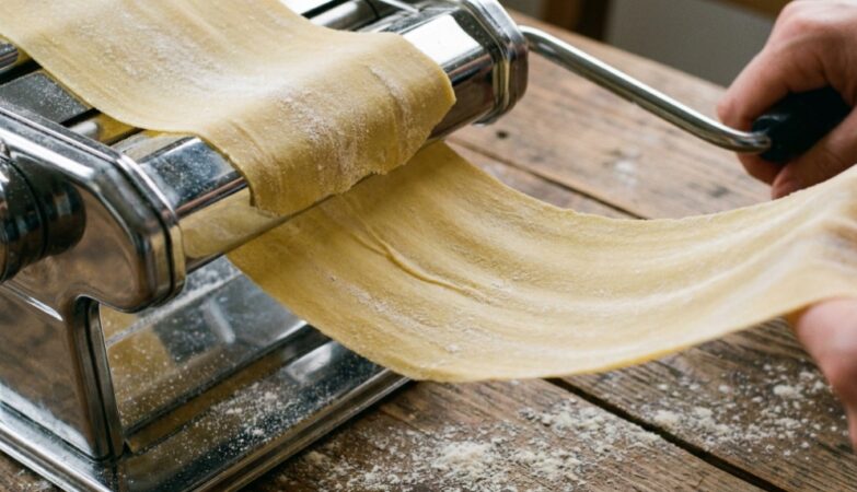 Hands adjusting the dial on a stainless steel pasta maker on a flour-dusted counter.